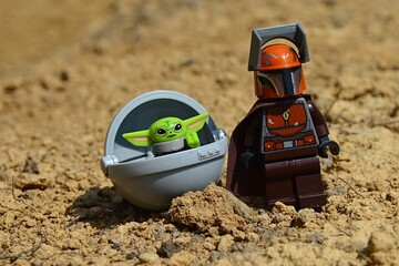 Naklejka premium LEGO Star Wars Mandalorian figure in brown suit with cloak standing on arid desert surface with alien Yoda race Child called Grogu in his flying pod, looking forward. 