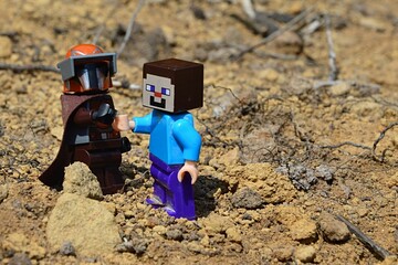 Fototapeta premium LEGO Mandalorian greets LEGO Minecraft Steve figure on arid desert like soil, summer daylight sunshine.