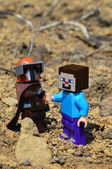 Fototapeta premium LEGO Mandalorian figure shaking hands with LEGO Minecraft Steve figure on arid desert like soil, summer daylight sunshine.