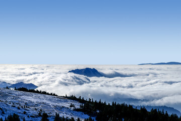 Fototapeta na wymiar An epic ocean of clouds and fog in the winter mountains landscape, aerial view. Huge white clouds come in waves over the foggy valleys, mountain peaks rise over like islands. Dramatic overcast sky
