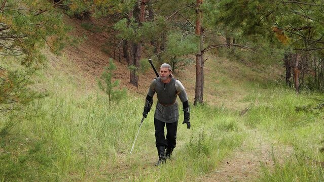 A medieval warrior with gray hair, dressed in chain mail with shoulder pads and a two-handed sword in his hands, carefully walks, crouching through the forest. Witcher, hunter of the Middle Ages