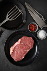 Entrecote Steak roh vom Rind mit Meersalz und roter Pfeffer auf Teller bei Vorbereitung in Küche mit Messer, Fleischgabel, Gusseisern Pfanne und Pfannenwender auf Schiefer Stein Hintergrund Schwarz