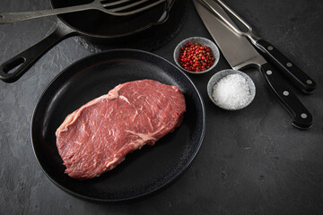 Entrecote Steak roh vom Rind mit Meersalz und roter Pfeffer auf Teller bei Vorbereitung in Küche mit Messer, Fleischgabel, Gusseisern Pfanne und Pfannenwender auf Schiefer Stein Hintergrund Schwarz