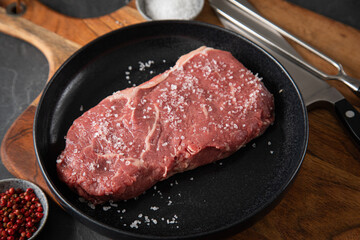 Entrecote Steak roh vom Rind mit Meersalz und roter Pfeffer auf Teller bei Vorbereitung in Küche mit Messer, Fleischgabel und Holz Brett auf Schiefer Stein Hintergrund Schwarz