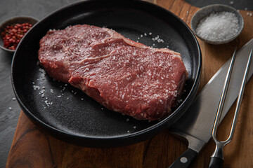 Entrecote Steak roh vom Rind mit Meersalz und roter Pfeffer auf Teller bei Vorbereitung in Küche mit Messer, Fleischgabel und Holz Brett auf Schiefer Stein Hintergrund Schwarz