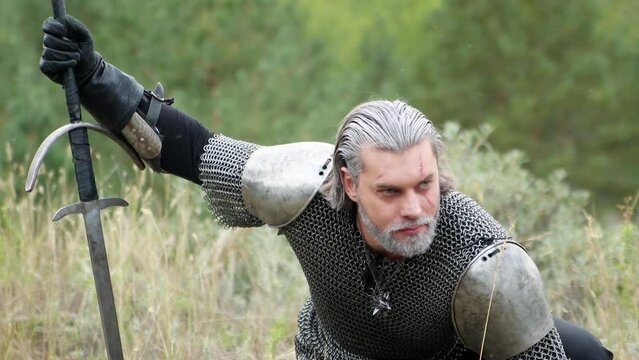 Smooth camera movement, sharp hay focus from bokeh to a warrior in chain mail with gray hair with a scar on his face and a two-handed sword in the hand of a sitting looking for traces. Hunter witcher