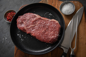 Entrecote Steak roh vom Rind mit Meersalz und roter Pfeffer auf Teller bei Vorbereitung in Küche mit Messer, Fleischgabel und Holz Brett auf Schiefer Stein Hintergrund Schwarz