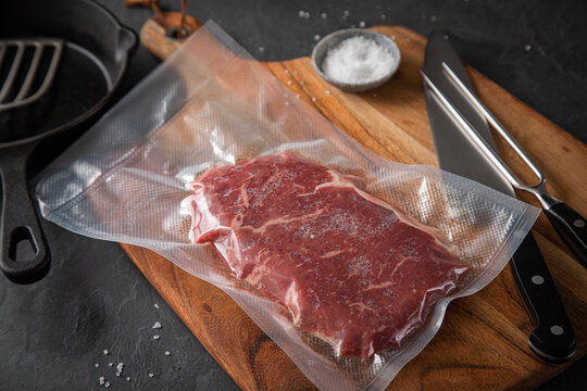 Entrecote Steak roh vakuumiert vom Rind mit Meersalz auf Teller bei Vorbereitung in K&uuml;che mit Messer, Fleischgabel, Gusseisern Pfanne, Plastik Vakuumbeutel und Pfannenwender auf Holzbrett und Schiefer