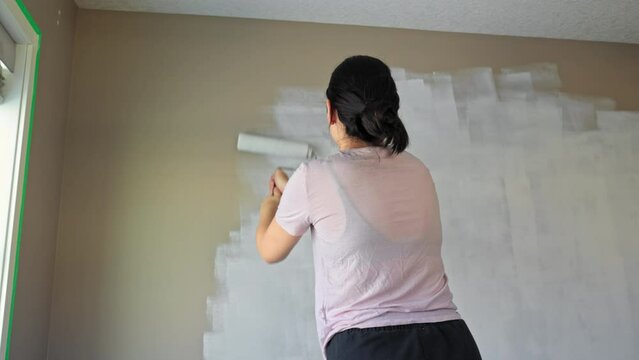 White woman painting a wall white near a window with a paint roller. 