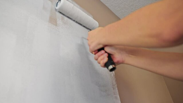 Close up of a caucasian person painting a wall white with a paint roller using both hands.