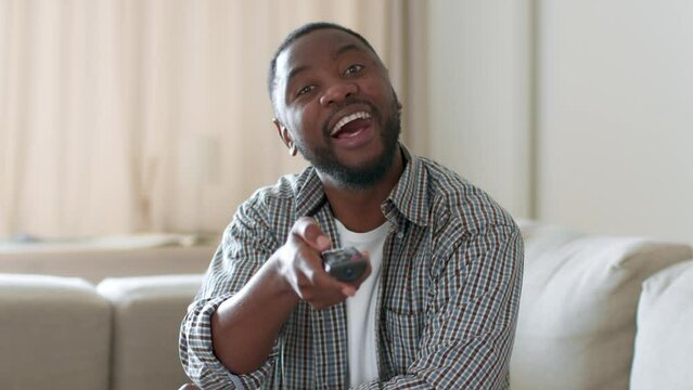 Funny tv content. Young interested african american man switching channels on tv with remote controller, free space