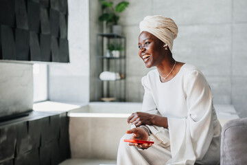 Cheerful African woman in white dress and turban holds phone sitting at home on couch. Happy...