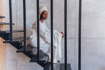 Confident African young woman in white turban and white dress sits on stairs at home looks at...