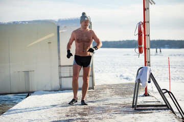 A man in warm hat and gloves swimming in an ice hole, walking along the pier. Winter activities in...