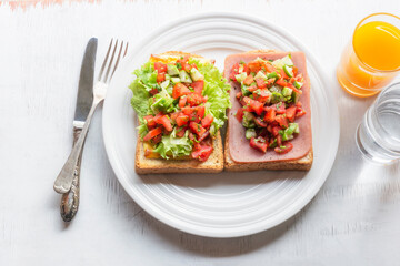 Sandwich breakfast has ham cheese and tomato cucumber on white wood table.