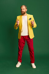 Portrait of stylish mature man in bright jacket and trousers isolated over green background. Concetp of fashion, beauty, emotions