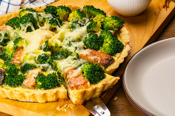 Freshly baked broccoli and salmon homemade quiche in black tart pan on metal cooling rack with moka pot coffee maker on wooden table.