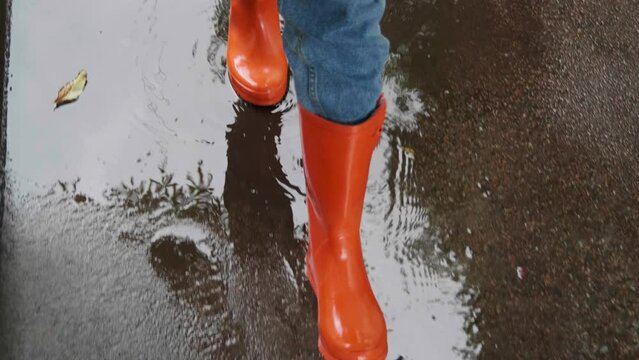 Woman in bright orange rain boots walking through puddles outdoors, above closeup view. Camera moving backwards