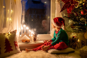 Toddler child, cute blond boy, sitting on the window in pajama, looking out for Satna Claus, Christmas lights around him