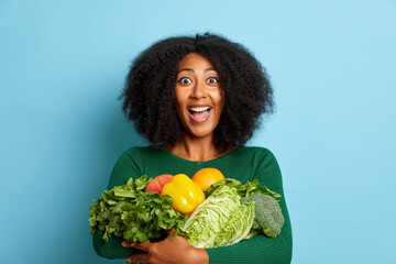 Woman holds a lot of vegetables, she likes healthy lifestyle and nutrition, so she smiles happily...