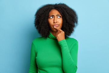 Curly haired woman in thinking pose, put finger to mouth and looks a side, isolated against blue background