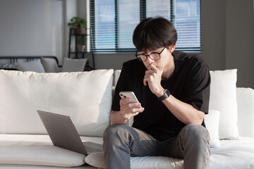 Portrait of an attractive serious young university student man man wearing casual clothes sitting on a couch at the living room, checking mail on mobile phone while using laptop.