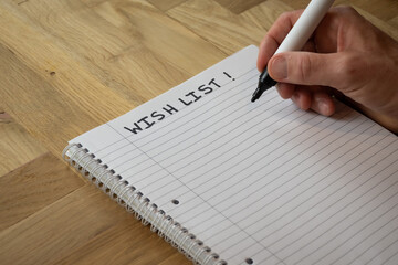 man writing a wishlist on plain lined notepad paper on a wooden desk where a wish list of things...