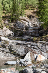 Granite karst rocks once washed by the Lillaz waterfall (Cascate di Lillaz) stream, and now dry under the midday sun, surrounded by slender trees, Aosta valley, Italy
