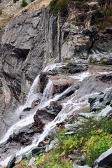 Foaming cascades of an alpine waterfall on granite rocks of various textures and colors overgrown with sparse green vegetation and mosses, vertical shot. Aosta valley, Italy