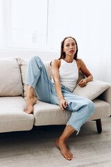 Obraz premium A beautiful young woman lies at home on the couch in a relaxed pose in a white tank top and blue jeans