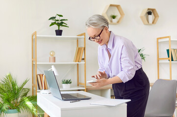 Joyful senior business woman in her office uses smartphone to organize business schedule. Stylish gray-haired woman near her workplace with laptop, who solves business cases with help of mobile phone.