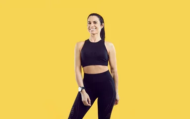 Foto op Canvas Portrait of beautiful happy successful female sports trainer isolated on yellow background. Happy millennial woman with athletic figure dressed in black in leggings and top smiling at camera. Banner. © Studio Romantic