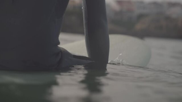 Close-up shot Surfer wearing wetsuit sits on his board, Water surface level Slow motion shot