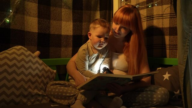 A young pregnant mother and a little son in pajamas in a bed house read a book and shine a flashlight. In the background is a Christmas garland. High quality 4k footage