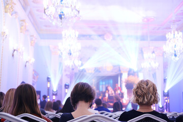 Guests in evening dresses before the start of the event sit in the hall illuminated by spotlights.