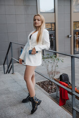 Blond stylish woman at oversized white jacket and phone on hand posing at the railing and steps on the city background