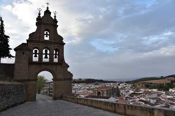 Classic Spanish triple bell tower of Iglesia Prior de Nuestra Señora del Mayor Dolor church, the white Andalusian town of Aracena and pastel blue, partly cloudy sky in the back, Aracena, Spain