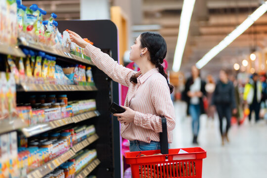 Side view of young Caucasian woman reaches hand to top shelf, holds cellphone and cart. Shelves with food in background. Concept of shopping in grocery store