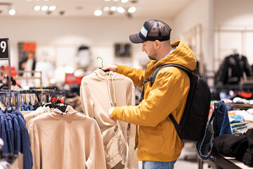 Bearded Caucasian young man wearing backpack chooses clothes in store. Concept of shopping and...
