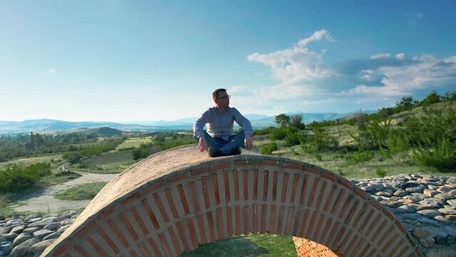Aerial view of man doing yoga exercise sitting on archaeological site arch at sunset