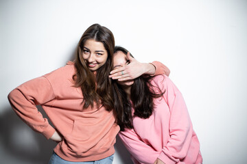 Two young attractive brunette girls in pink sweaters embracing each other smiling, one is closing the eyes of another with her hand, white background