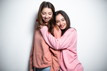 Two young attractive brunette girls in pink sweaters embracing each other, standing one after another, smiling, looking at the camera, white background