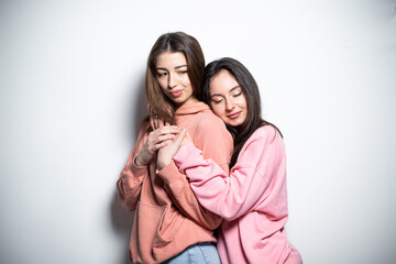 Two young attractive brunette girls in pink sweaters embracing each other, standing one after another with closed eyes, smiling, looking at the camera, white background