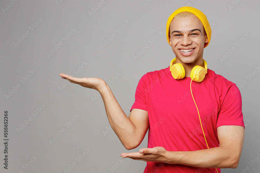 Wall mural Young smiling happy fun man of African American ethnicity 20s he wearing pink t-shirt yellow hat headphones point hands arms aside on workspace area mock up isolated on plain grey background studio.