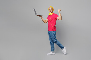 Full body fun side view young man of African American ethnicity wear pink t-shirt yellow hat headphones hold use work on laptop pc computer do winner gesture isolated on plain grey background studio.