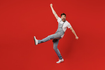 Fototapeta na wymiar Full body side view overjoyed young male housewife housekeeper chef cook baker man wear grey apron raise up hands do winner gesture clench fists isolated on plain red background. Cooking food concept.