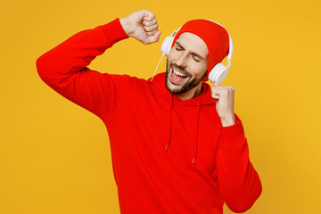 Young smiling happy fun caucasian man wearing red hoody hat headphones listening to music gesticulating with hands isolated on plain yellow color background studio portrait. People lifestyle concept.