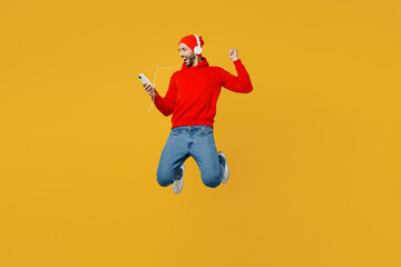 Obraz premium Full body young fun man wear red hoody hat headphones listen to music jump high hold in hand use mobile cell phone isolated on plain yellow color background studio portrait. People lifestyle concept.