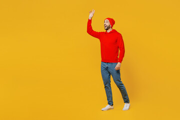 Full body side view smiling happy fun young caucasian man wears red hoody hat walking going strolling waving hand isolated on plain yellow color background studio portrait. People lifestyle concept.