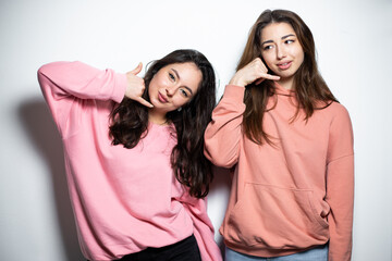 Two young attractive brunette girls in pink sweaters , smiling, showing call me gesture, white background, call center 